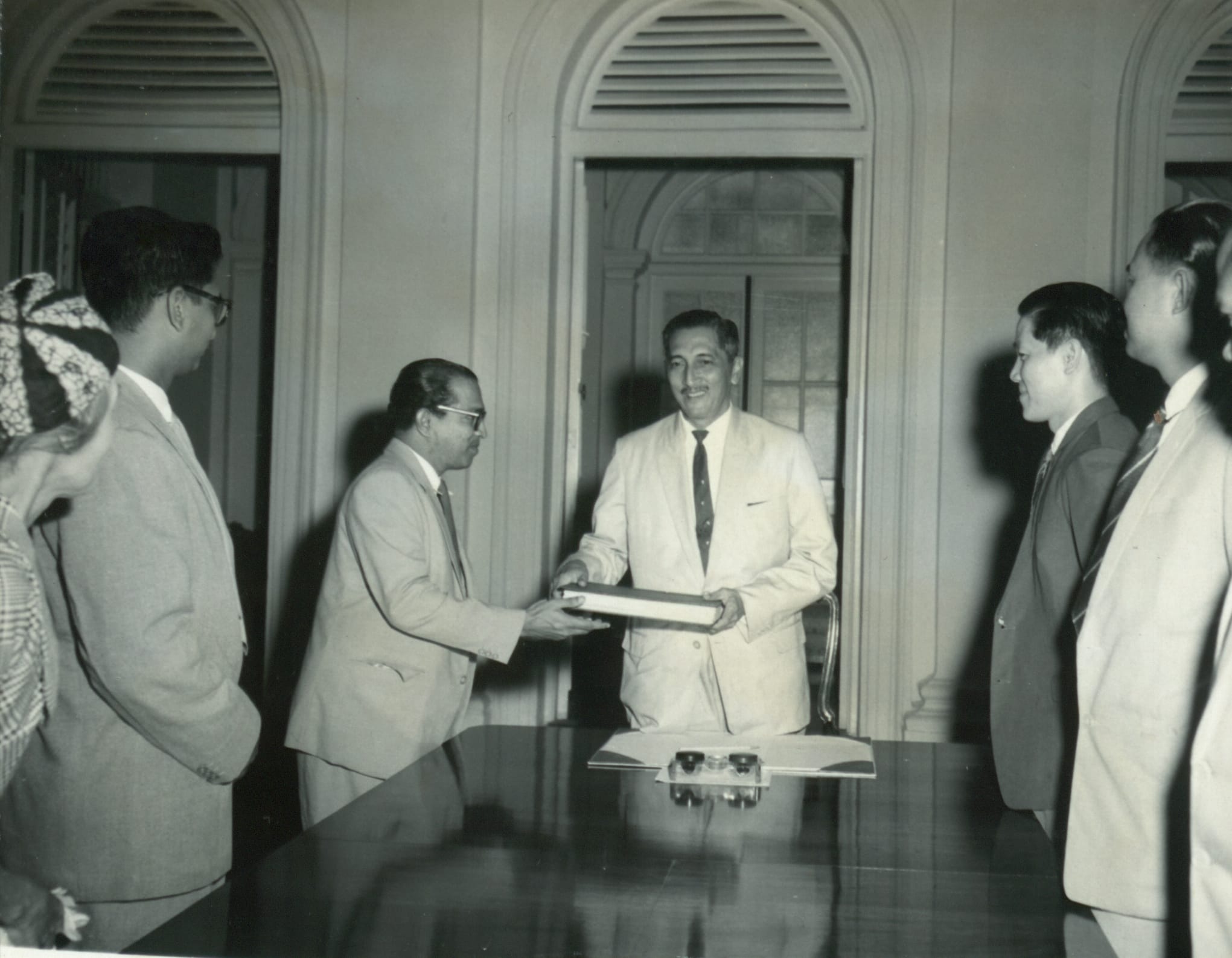 Several men in suits stand around a table, with two men exchanging a document in a room with arched windows.